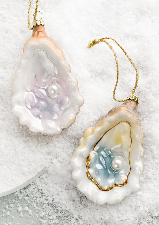 Two glass holiday ornaments of oyster half shell with pearl, one is blue and one is lavender.
